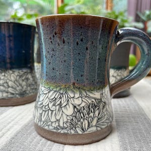 May include: Handmade ceramic mug with a floral design. The mug features a dark blue and brown glaze with a white floral pattern on the lower half. The handle is a dark blue-green color. The mug is suitable for hot or cold beverages.
