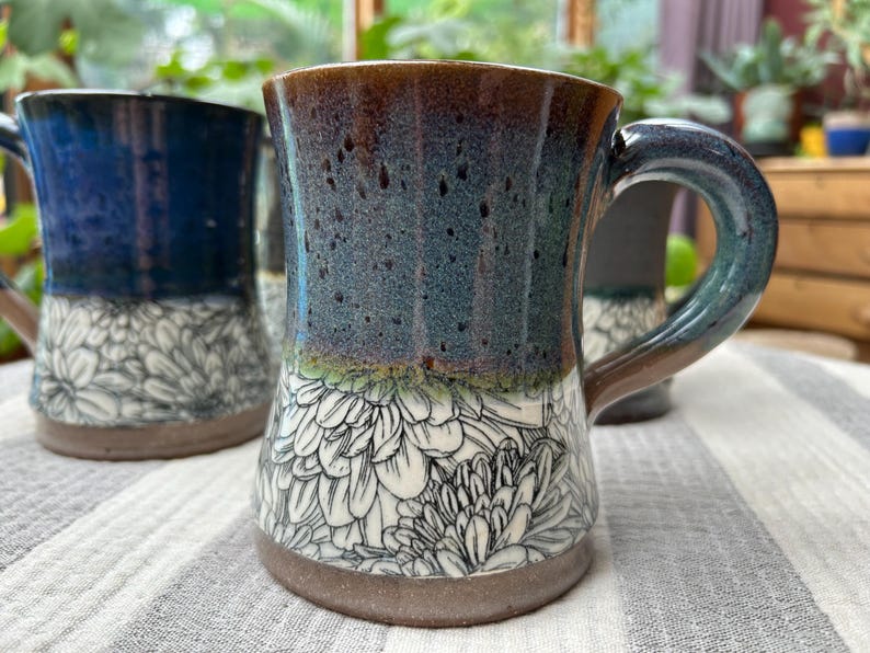 May include: Handmade ceramic mug with a floral design. The mug features a dark blue and brown glaze with a white floral pattern on the lower half. The handle is a dark blue-green color. The mug is suitable for hot or cold beverages.