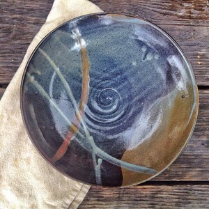 May include: A large, round ceramic plate with a blue, brown, and white glaze. The plate has a swirl design in the center and a variety of abstract lines and shapes around the edge.