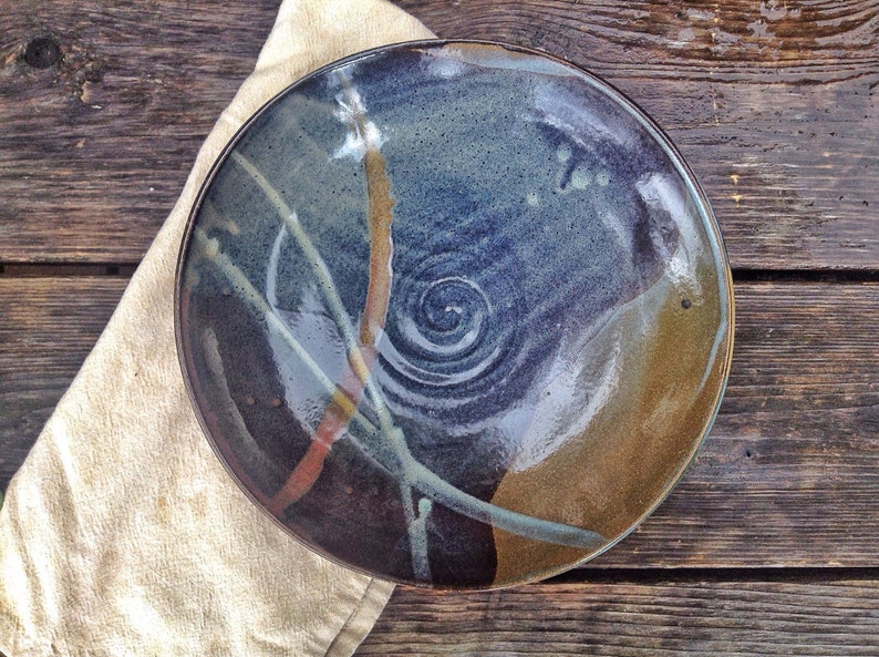 May include: A large, round ceramic plate with a blue, brown, and white glaze. The plate has a swirl design in the center and a variety of abstract lines and shapes around the edge.