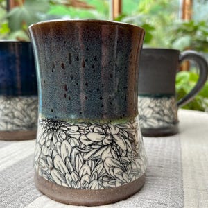 May include: Three ceramic mugs with a floral design. The mugs have a dark blue, speckled glaze at the top, transitioning to a white floral pattern on the bottom. The mugs are on a striped surface.