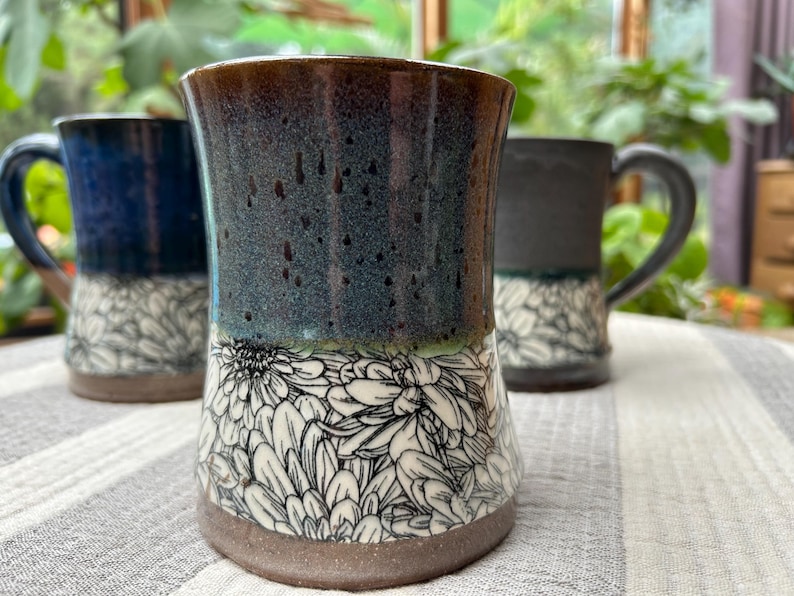 May include: Three ceramic mugs with a floral design. The mugs have a dark blue, speckled glaze at the top, transitioning to a white floral pattern on the bottom. The mugs are on a striped surface.