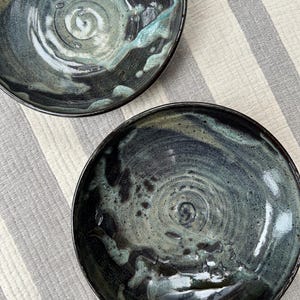 Puede incluir: Dos cuencos de cer&aacute;mica hechos a mano con un acabado oscuro y brillante. Los cuencos presentan un patr&oacute;n arremolinado de color turquesa, gris y negro, con un dise&ntilde;o en espiral en el centro. Los cuencos est&aacute;n sobre un fondo a rayas.