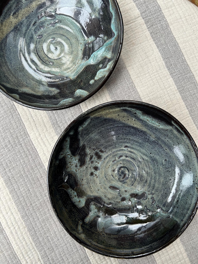 Puede incluir: Dos cuencos de cer&aacute;mica hechos a mano con un acabado oscuro y brillante. Los cuencos presentan un patr&oacute;n arremolinado de color turquesa, gris y negro, con un dise&ntilde;o en espiral en el centro. Los cuencos est&aacute;n sobre un fondo a rayas.