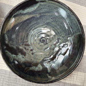 Puede incluir: Un cuenco de cer&aacute;mica hecho a mano con un acabado oscuro y brillante. El cuenco presenta una mezcla de tonos azul oscuro, verde y marr&oacute;n, con un patr&oacute;n en espiral en el centro. El borde es oscuro y el interior tiene un aspecto texturizado.