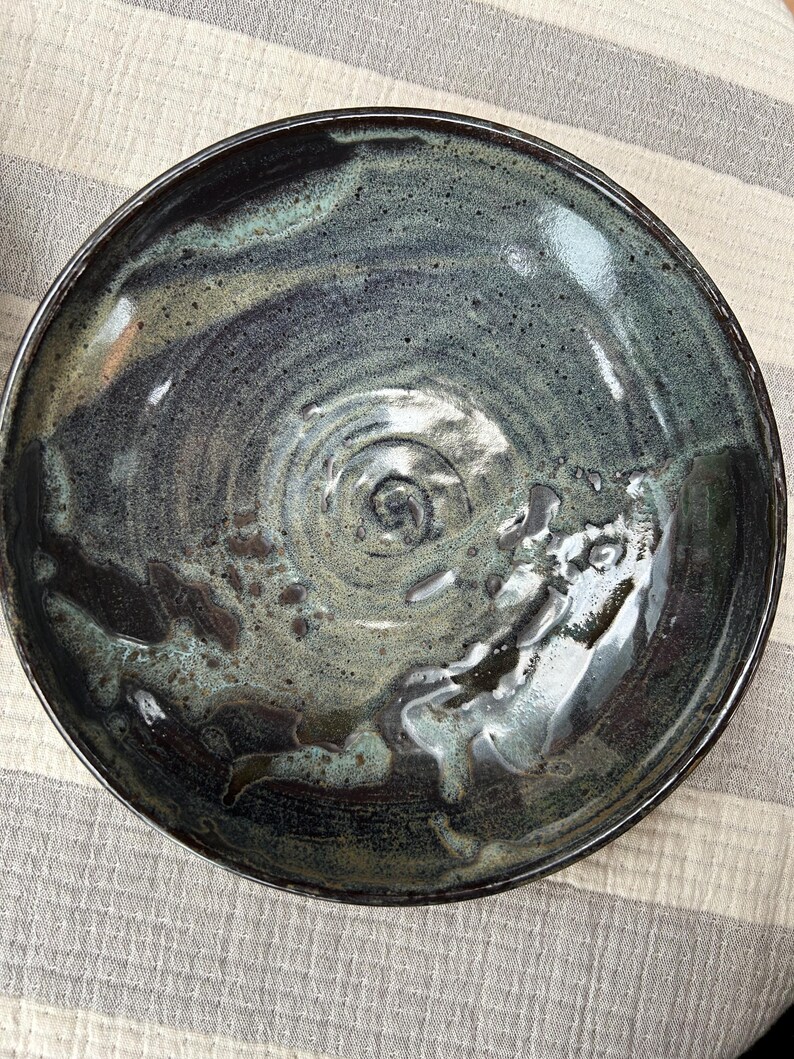 Puede incluir: Un cuenco de cer&aacute;mica hecho a mano con un acabado oscuro y brillante. El cuenco presenta una mezcla de tonos azul oscuro, verde y marr&oacute;n, con un patr&oacute;n en espiral en el centro. El borde es oscuro y el interior tiene un aspecto texturizado.