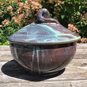 May include: A handcrafted ceramic lidded bowl with a rich, earthy brown base and a lid with teal and blue marbling. The lid features a decorative swirl handle. The bowl is set on a wooden surface, showcasing its unique, artistic design.