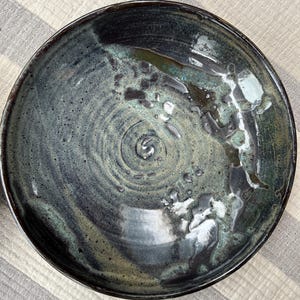May include: A handmade ceramic bowl with a dark blue and teal glaze. The bowl has a wide, shallow shape with a textured surface and a spiral design in the center. The rim is dark, and the interior has a glossy finish.
