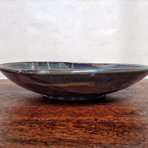 May include: A dark blue ceramic bowl with a wide rim and a slightly raised base. The bowl has a mottled glaze with streaks of brown, white, and green.