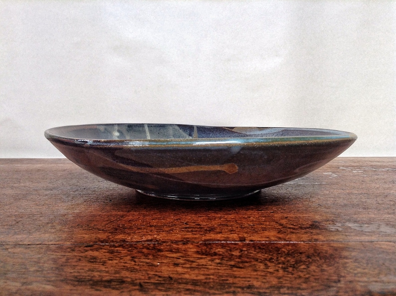 May include: A dark blue ceramic bowl with a wide rim and a slightly raised base. The bowl has a mottled glaze with streaks of brown, white, and green.