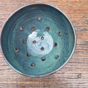 May include: A teal blue ceramic colander with a speckled glaze and a round shape. The colander has a series of small holes throughout the bottom.