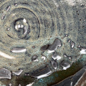 Puede incluir: Primer plano de un plato de cer&aacute;mica con superficie texturizada. El plato presenta un dise&ntilde;o en espiral en tonos grises y verde azulado, con acentos blancos y grises. El esmalte tiene un aspecto moteado.