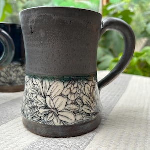 May include: A handmade ceramic mug with a dark gray upper section and a white floral pattern on the lower half. The mug has a curved handle and a dark gray base. The background includes other mugs and greenery.