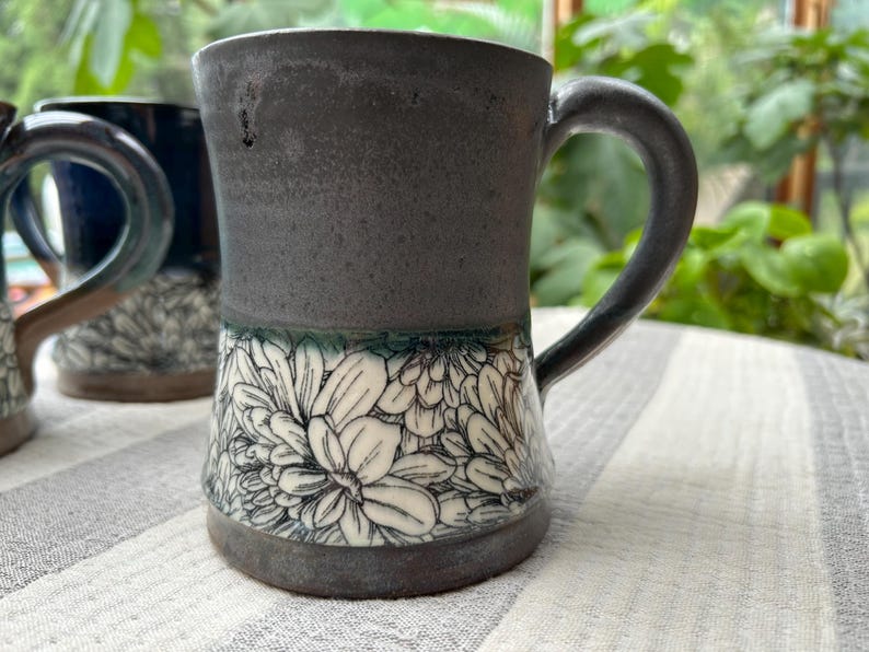 May include: A handmade ceramic mug with a dark gray upper section and a white floral pattern on the lower half. The mug has a curved handle and a dark gray base. The background includes other mugs and greenery.