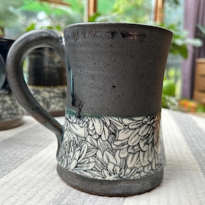 May include: A handmade ceramic mug with a dark gray exterior and a white floral pattern on the lower half. The mug has a large handle and is sitting on a striped surface.