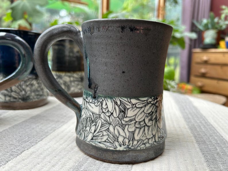May include: A handmade ceramic mug with a dark gray exterior and a white floral pattern on the lower half. The mug has a large handle and is sitting on a striped surface.