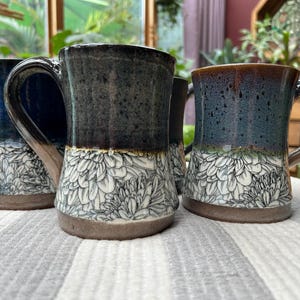 May include: Four ceramic mugs with a floral design. The mugs have a dark blue, teal, and brown glaze. The bottom half of each mug features a white floral pattern. Each mug has a large handle and a brown base.