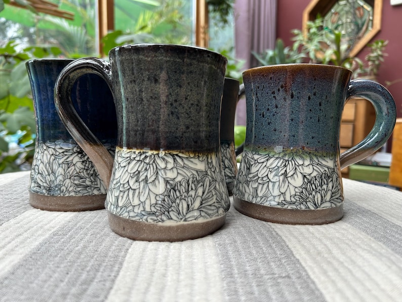May include: Four ceramic mugs with a floral design. The mugs have a dark blue, teal, and brown glaze. The bottom half of each mug features a white floral pattern. Each mug has a large handle and a brown base.