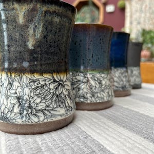 May include: A row of handmade ceramic tumblers with a floral design. The tumblers have a dark, textured glaze on the top half and a white floral pattern on the bottom half. The colors include dark blue, black, and white.