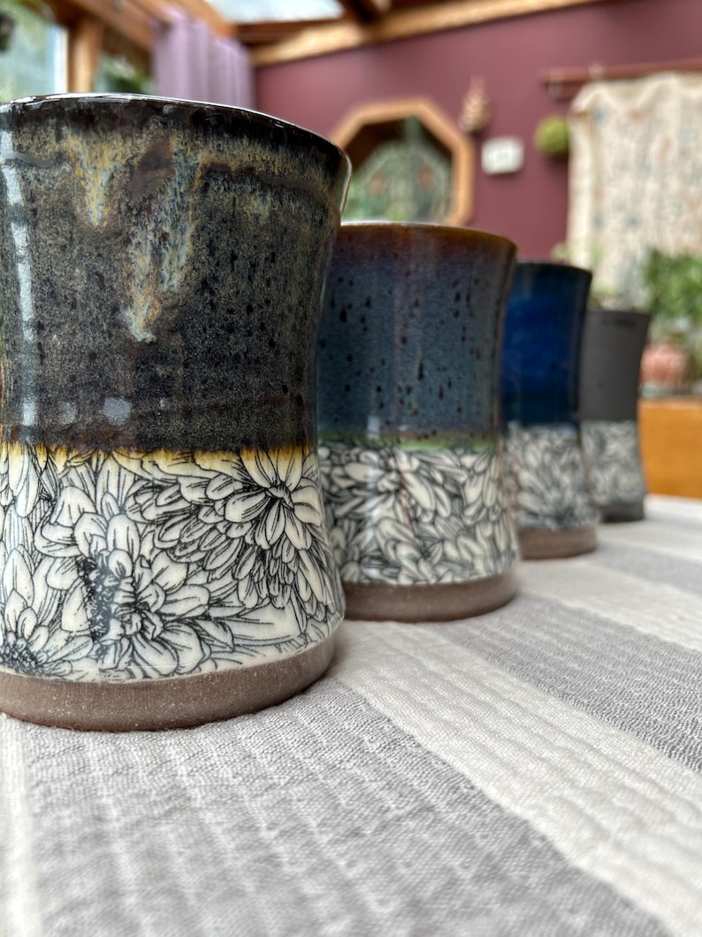 May include: A row of handmade ceramic tumblers with a floral design. The tumblers have a dark, textured glaze on the top half and a white floral pattern on the bottom half. The colors include dark blue, black, and white.