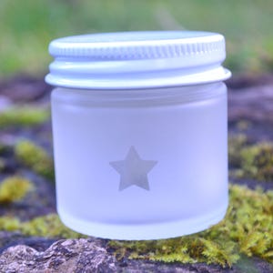 May include: A small, frosted glass jar with a silver lid. A gray star is etched on the front of the jar. The jar is sitting on a mossy, natural surface.