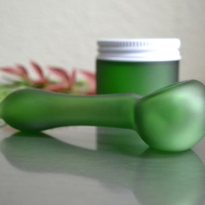 May include: A frosted green glass pipe with a rounded bowl. The pipe is lying on a reflective surface.