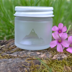 May include: A frosted glass jar with a white lid and a mountain design. The jar is sitting on a mossy log with three pink flowers.