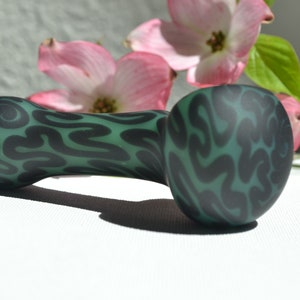 May include: A green glass pipe with a black swirl design. The pipe is on a white surface with pink flowers in the background.