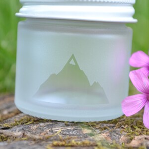 May include: A frosted glass jar with a white lid and a mountain silhouette etched into the side. The jar is sitting on a piece of wood with pink flowers in the background.
