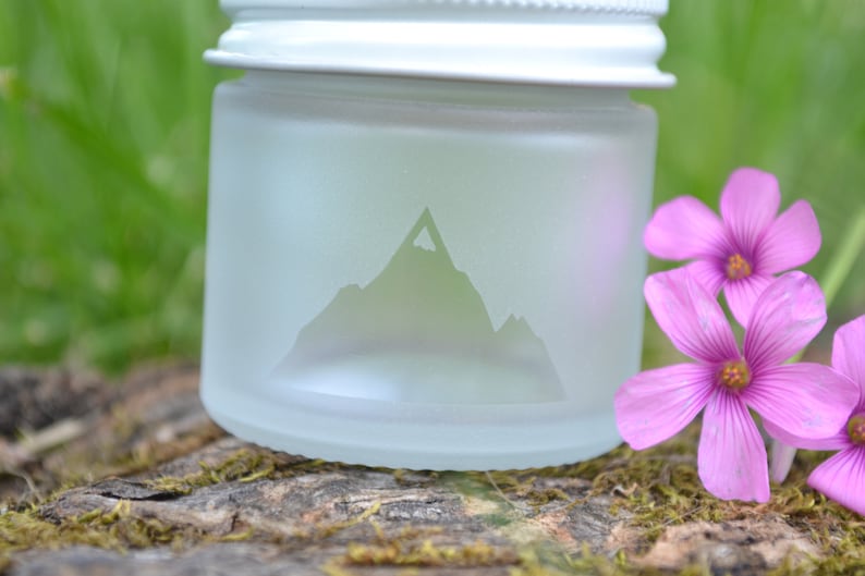 May include: A frosted glass jar with a white lid and a mountain silhouette etched into the side. The jar is sitting on a piece of wood with pink flowers in the background.