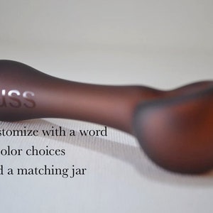 May include: A frosted glass pipe with the word "Russ" etched into the side. The pipe is a dark brown color. The text "Customize with a word", "7 Color choices", and "Add a matching jar" is visible below the pipe.