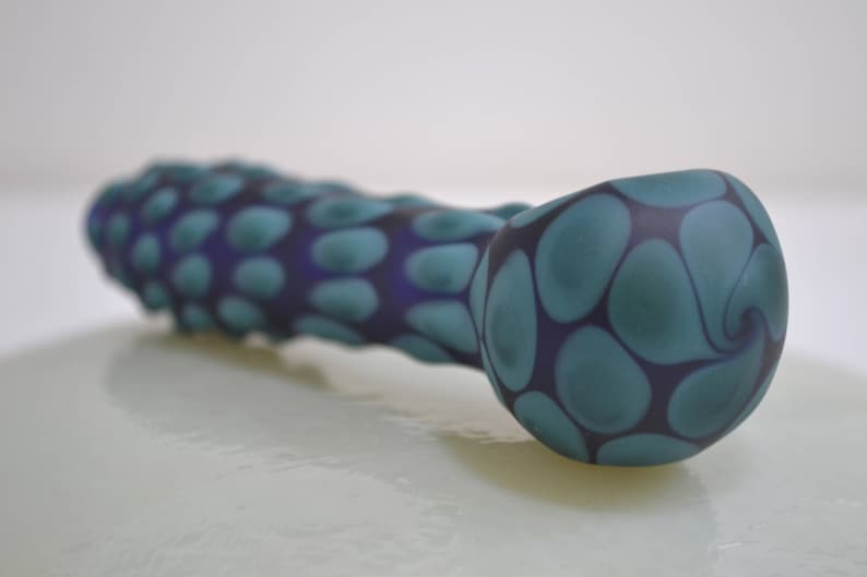 Op de afbeelding: Een blauwe en paarse glazen pijp met een draaiend patroon. De pijp is gevormd als een lange, dunne buis met een afgeronde kom aan het ene uiteinde.