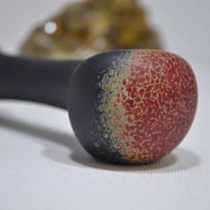 May include: A black glass pipe with a red and black speckled design. The pipe has a round bowl and a long, thin stem.