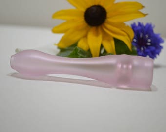 May include: A light pink glass bottle stopper with a rounded shape. A yellow sunflower and a blue flower are in the background. The stopper is approximately 6 inches long.