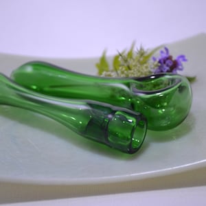 May include: Two green glass smoking pipes on a white plate. The pipes are made of clear glass with a green tint. The pipes are lying on their sides, with the bowl of the pipe facing up.