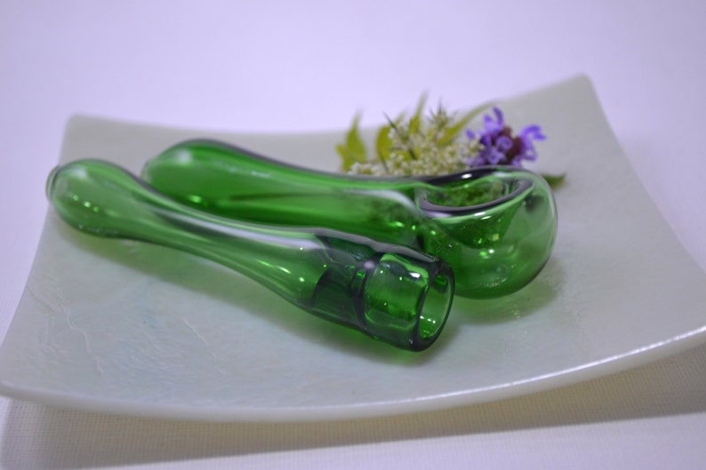 May include: Two green glass smoking pipes on a white plate. The pipes are made of clear glass with a green tint. The pipes are lying on their sides, with the bowl of the pipe facing up.