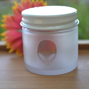 Puede incluir: Un tarro de vidrio esmerilado con tapa blanca y un gr&aacute;fico de cara de extraterrestre. El tarro est&aacute; sobre una superficie de madera.