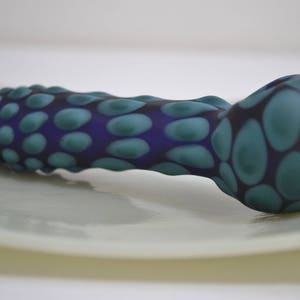 Op de afbeelding: Een blauwe en groene glazen pijp met een getextureerd, hobbelig oppervlak. De pijp is gevormd als een lange, dunne buis met een afgeronde kom aan het ene uiteinde.