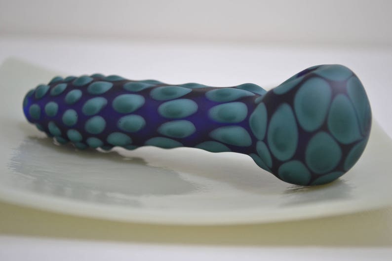 Op de afbeelding: Een blauwe en groene glazen pijp met een getextureerd, hobbelig oppervlak. De pijp is gevormd als een lange, dunne buis met een afgeronde kom aan het ene uiteinde.