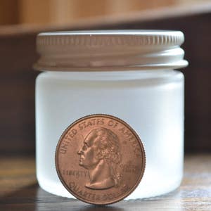 Puede incluir: Un tarro de vidrio esmerilado con tapa blanca. El tarro es del tama&ntilde;o aproximado de una moneda de 1 euro.