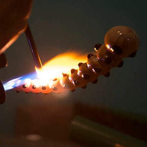 May include: A glass pipe being heated with a torch. The pipe is shaped like a caterpillar with black dots and a light brown body.