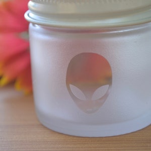 Puede incluir: Un tarro de vidrio esmerilado con tapa blanca y una cara de extraterrestre grabada en la parte delantera.