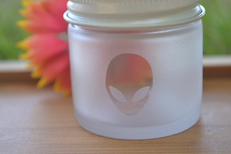 Puede incluir: Un tarro de vidrio esmerilado con tapa blanca y una cara de extraterrestre grabada en la parte delantera.