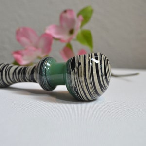 May include: A black and white striped glass pipe with a green accent. The pipe is lying on a white surface.