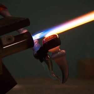 May include: A glassblowing torch with a blue and orange flame is heating a clear glass pipe with a white tip. The pipe is being held in place by a metal clamp.