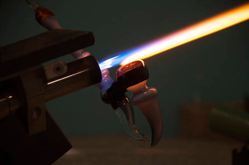 May include: A glassblowing torch with a blue and orange flame is heating a clear glass pipe with a white tip. The pipe is being held in place by a metal clamp.
