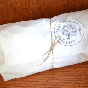 Op de afbeelding: Een wit papieren pakketje vastgebonden met touw en een blauwe stempel met de tekst "Lone Oak Glassworks L.O. Glass".
