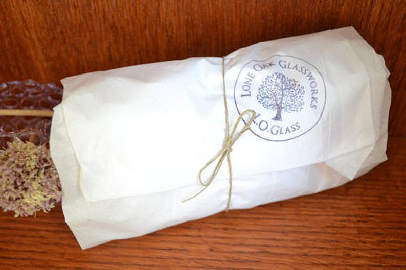 Op de afbeelding: Een wit papieren pakketje vastgebonden met touw en een blauwe stempel met de tekst "Lone Oak Glassworks L.O. Glass".