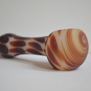 May include: A white glass pipe with brown and white swirl patterns. The pipe is shaped like a long, thin tube with a rounded end.