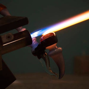 Op de afbeelding: Een glasblaasbrander met een blauwe en oranje vlam verhit een glazen pijp. De pijp wordt gevormd tot een gebogen ontwerp.